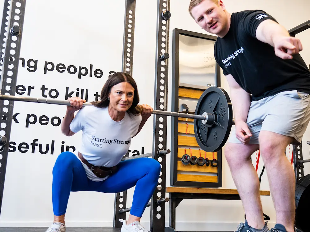 Female trainee in a Starting Strength Gym performing a squat while male Coach provides instruction
