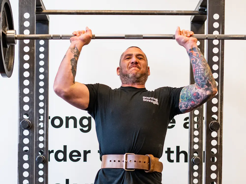 Male athlete wearing lifting belt performing overhead press at Starting Strength gym